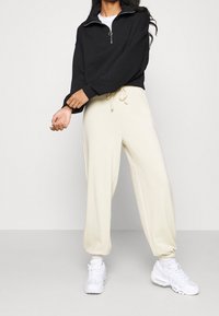 Sweatshirt noir court avec fermeture éclair quart, col rond, associé à un pantalon de jogging crème à taille élastique, complété par des baskets blanches.