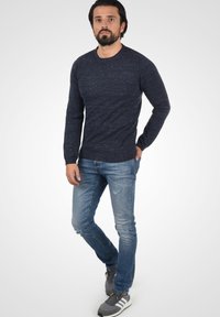 Sötétkék kötött pulóver, kerek nyakkal, világoskék, koptatott farmernadrággal és szürke sportcipővel, fehér csíkkal viselve.