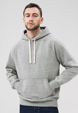 Mann mit kurzem Haar und Bart trägt einen hellgrauen Hoodie mit Fronttasche und weißen Kordeln und steht vor einem schlichten Hintergrund.