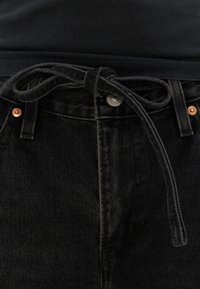 Jean en denim noir avec une ceinture en tissu nouée à la taille, des rivets en cuivre et un haut noir rentré à l'intérieur.