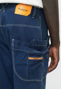 Ναυτικές παντελόνες εργασίας με ενισχυμένες ραφές, προεξοχές τσέπες και μια πορτοκαλί δερμάτινη ετικέτα που φέρει το "Replay Inc." στη ζώνη της πλάτης.