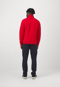Sudadera roja con cuello alto, combinada con pantalones de chándal en azul marino oscuro. La tela parece suave, con un corte relajado y mínimos detalles de diseño.