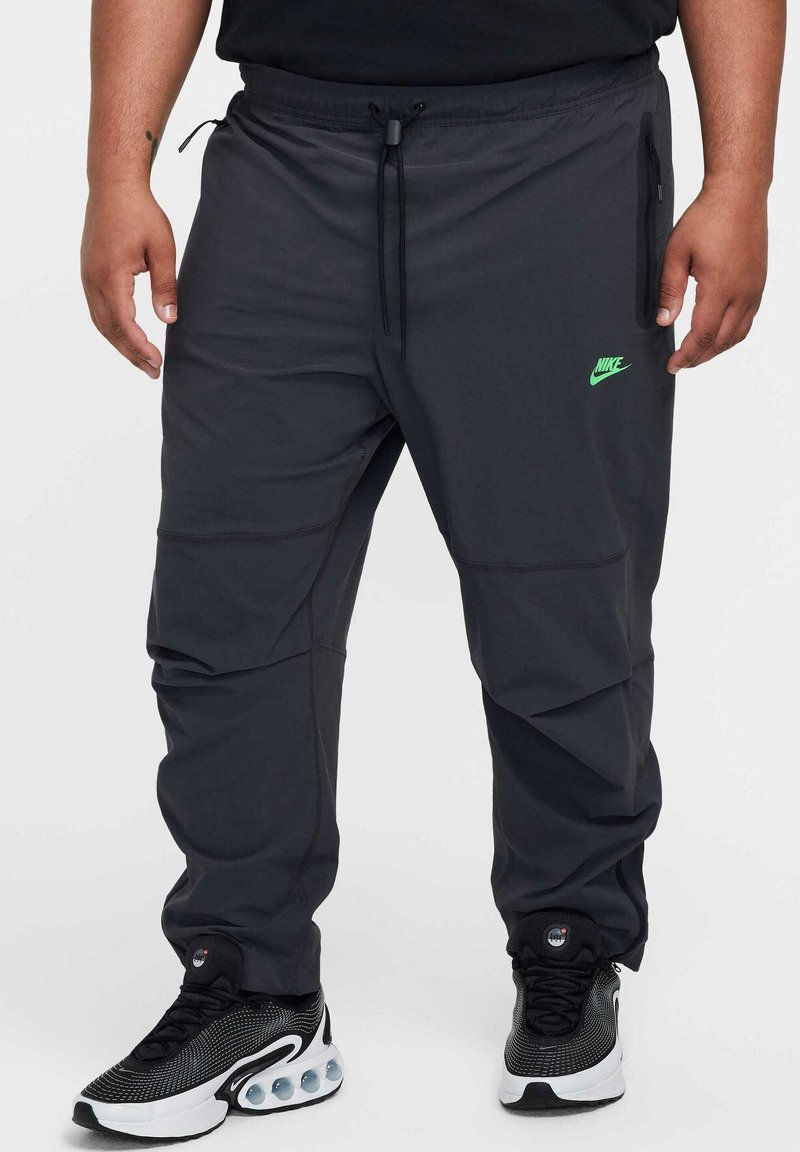 Pantaloni atletici grigi realizzati in tessuto leggero, dotati di una cintura regolabile, tasche laterali e di un logo Nike verde sulla coscia.