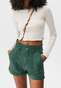 Mujer con una camiseta corta de manga larga de canalé color crema, con cortes en forma de botón en diagonal, y pantalones cortos verdes de cintura alta con dobladillo enrollado y bolsillos.