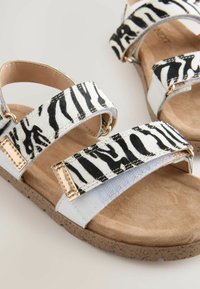 Sandali con stampa zebra bianca e nera, dotati di due cinturini regolabili, con dettagli dorati e una soletta texturizzata color beige. Suola in gomma con dettagli maculati.