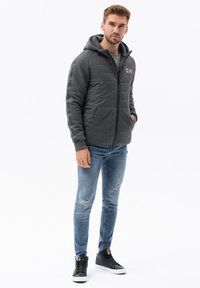 Chaqueta gris con capucha, parte frontal acolchada y dos bolsillos delanteros. Combinada con jeans azules desgastados y zapatillas altas negras.