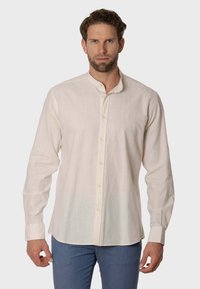 Camisa de manga larga color beige claro, confeccionada en una tela liviana, que presenta un cuello mao y un sutil logo bordado en el pecho.