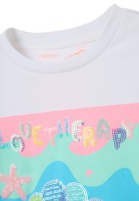 Camiseta de algodón blanca con escote redondo que presenta texto en lentejuelas multicolores "LOVETHERAPY" y patrones de olas en rosa y azul.