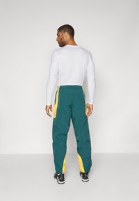 Pantaloni sportivi verdi con strisce laterali gialle e vestibilità rilassata. Tessuto leggero con una texture liscia. Sneakers nere.