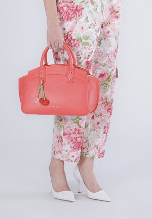 Osoba v květovaných kalhotách a bílých podpatcích držící korálově růžovou kabelku s přívěskem třešně na jednobarevném pozadí.