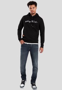 Sudadera negra con un logo en script "Wrong Friends", además de un bolsillo delantero. Combinada con jeans de mezclilla oscura y zapatillas negras y blancas.