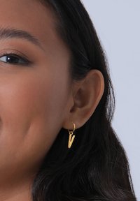 Goldene Creolen mit einem V-f örmigen Anhänger, strukturierten Oberflächen und einem schlichten Verschlussdesign. An dem Ohr eines Modells getragen, mit sichtbaren dunklen Haaren.