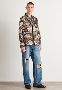 Camo-patterned shirt, cotton blend, long sleeves, button-up design. Worn with light blue distressed jeans, straight fit, frayed hems. Black shoes.