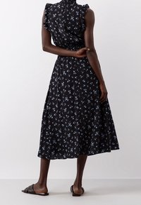 Robe noire en midi avec un motif floral bleu, manches volantées, corsage froncé et jupe fluide. Associée à des sandales glissantes noires simples.