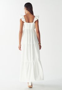 Valkoinen maksimekko, jossa on röyhelöolkaimet, smockattu selkä, kerroksellinen muotoilu ja virtaava helma. Materiaalina pehmeä, kevyt kangas ja rento istuvuus.