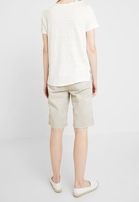 Valkoinen lyhythihainen t-paita, beiget shortsit ja valkoiset slip-on-kengät. T-paidassa on pieni sivuhalkio; shortseissa on takataskut.