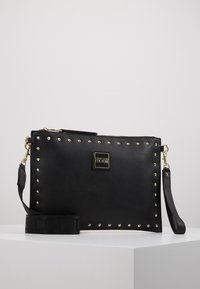 Borsa a tracolla in pelle nera con borchie dorate lungo i bordi, chiusura con zip e piastra con logo. Include una tracolla rimovibile con lettering in rilievo.