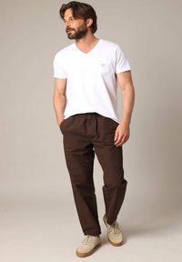 Witte v-hals t-shirt met een klein zakje, gecombineerd met bruine broek met elastische taille en beige veterschoenen op een neutrale achtergrond.