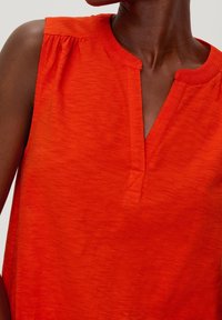 Červený top bez rukávů s výstřihem do V, na ramenou se sklady, měkká textura a volný střih. Materiál z bavlněné směsi.