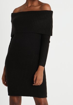 Femme portant une robe-pull noire côtelée à épaules dénudées avec manches longues, debout devant un fond blanc uni.