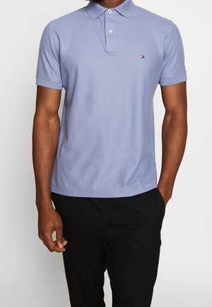 Homme portant un polo bleu clair à manches courtes avec un petit logo sur la poitrine et un pantalon noir, debout avec une main dans une poche.