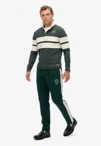 Dunkelgrüner Pullover mit weißen Streifen und Logo, kombiniert mit passenden grünen Hosen, die ebenfalls ein Logo tragen. Beide Artikel haben eine lässige Passform und Textur.