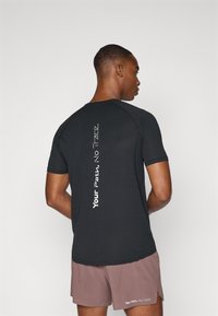 T-shirt de sport noir à manches courtes avec une finition texturée, avec un texte vertical blanc au dos. Associé à un short marron.