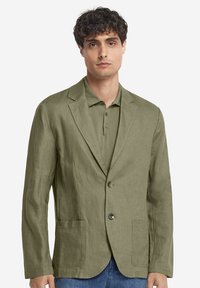 Olivgrüner Leinenblazer mit zwei Fronttaschen, einreihigem Knopfverschluss und lockerer Passform, getragen über einem passenden Poloshirt.
