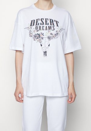 Personne portant un t-shirt blanc ample avec le texte "Desert Dreams" et un motif de crâne de longhorn fleuri, assorti à un pantalon blanc.