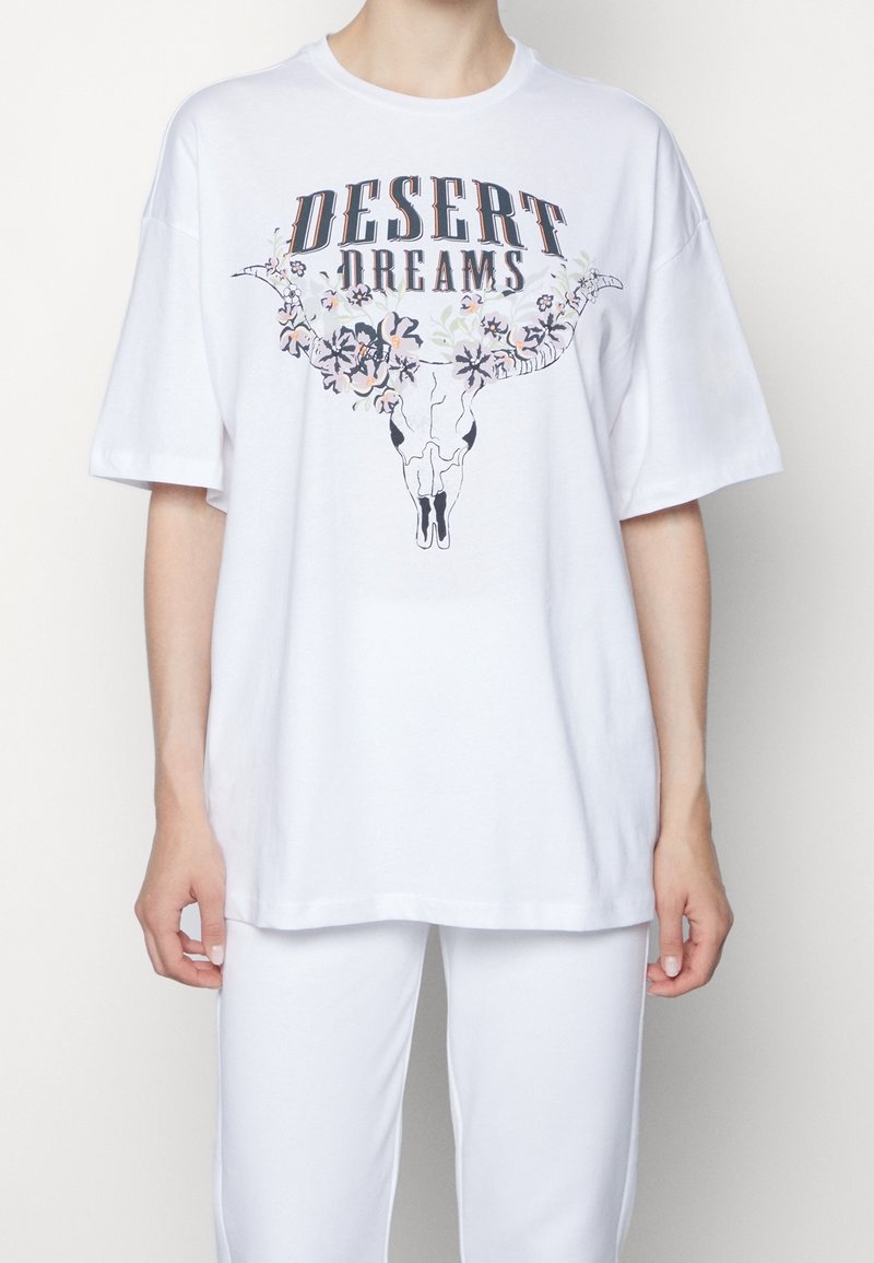 Personne portant un t-shirt blanc ample avec le texte "Desert Dreams" et un motif de crâne de longhorn fleuri, assorti à un pantalon blanc.
