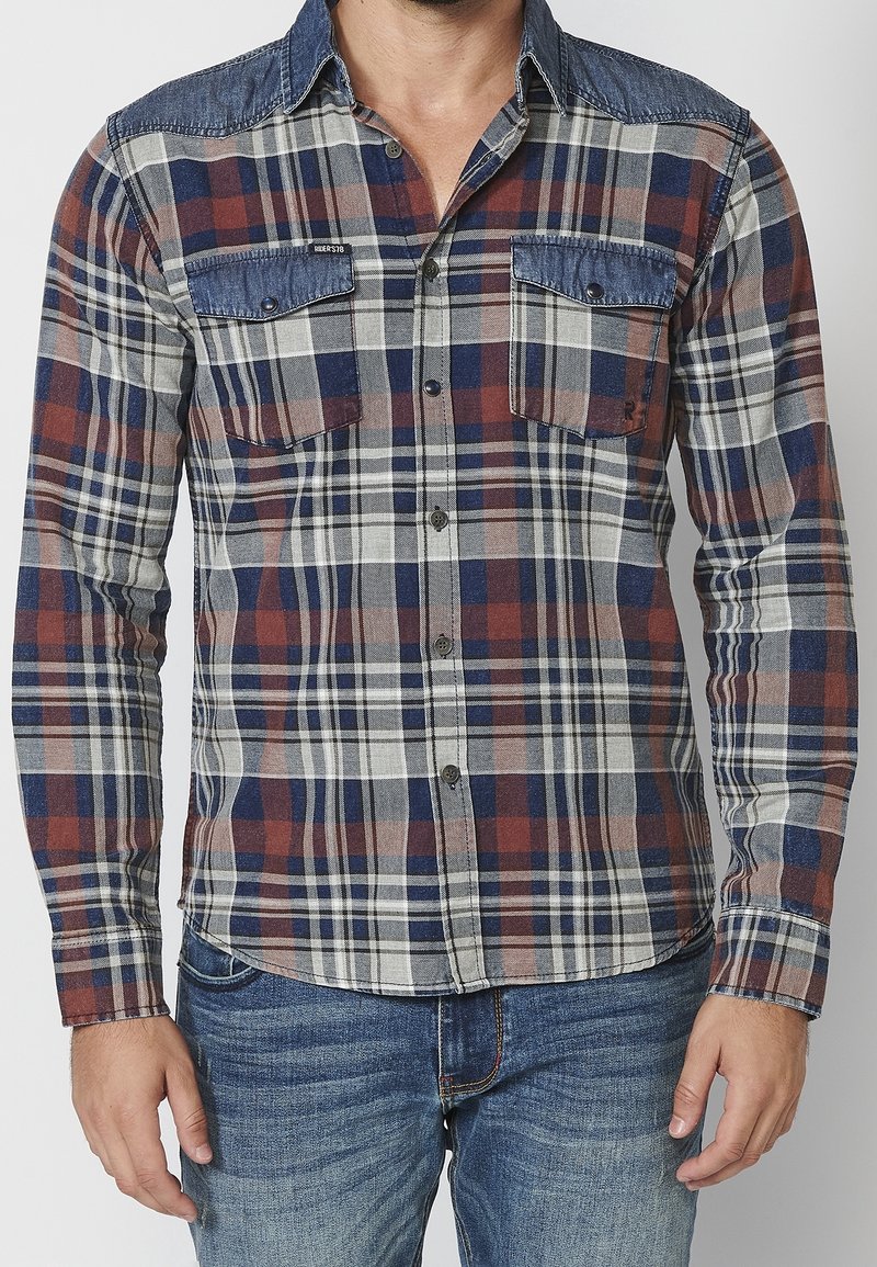 Homme portant une chemise à carreaux à manches longues avec des détails en denim bleu sur les poches et les épaules, associée à un jean bleu.