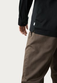 Schwarzes Langarmhemd mit Knopfmanschetten, kombiniert mit hellbraunen Hosen mit Taschen; sichtbares Markenlabel nahe dem Saum.