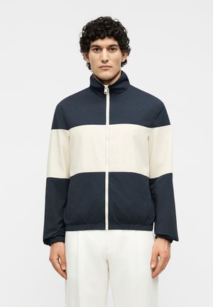 Άνδρας που φοράει ένα δίχρωμο (ασπρόμαυρο) μπουφάν με φερμουάρ, ελαστικά μανίκια και λευκό παντελόνι, στέκεται μπροστά σε απλό φόντο.