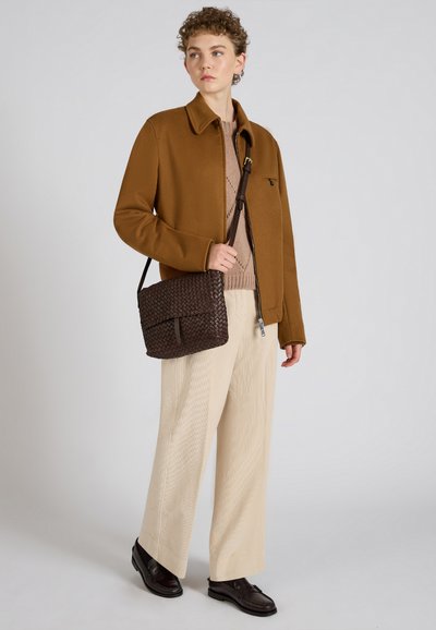 Bruine geweven schoudertas met flapsluiting, gedragen door een persoon in een bruine jas, beige trui en crèmekleurige wijde broek.