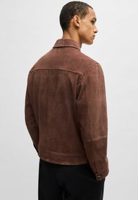 Giacca in suede marrone con colletto strutturato, tasche laterali e polsini con bottoni, caratterizzata da una texture liscia e linee pulite.