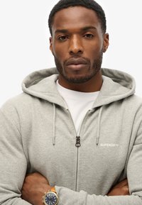 Grå zip-up hoodie med snore og et diskret logo. Har en blød tekstur, afslappet pasform og et hvidt underlag. Brun ur på håndleddet.