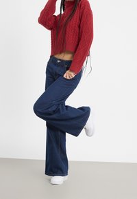 Czerwony sweter crop z dzianiny o splocie w warkocze, połączony z ciemnoniebieskimi spodniami typu wide-leg i białymi trampkami. Minimalna widoczna biżuteria.