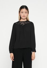 Svart bluse med lange ermer, et blonderpanel ved halslinjen og plisseringer. Stoffet har en glatt tekstur med samlete detaljer.