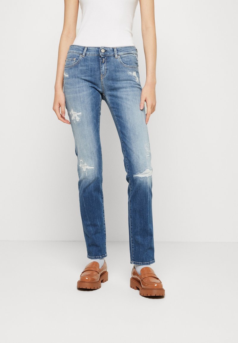 Jean denim bleu clair avec une coupe droite, présentant des accents déchirés et un léger dégradé. Associés à des mocassins marron pour créer un contraste.