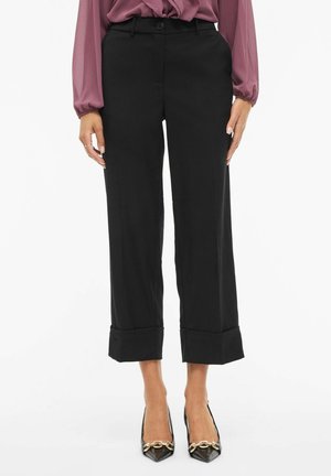 Svarte bukser med vide ben, glatt tekstur, knappelukking og oppbrettede mansjetter, kombinert med spisse svarte hæler med gullaksenter.
