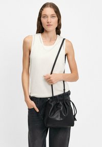 Schwarze Leder-Einkaufstasche mit Kordelzugverschluss, einem Schulterriemen und weicher Textur, kombiniert mit einem weißen ärmellosen Oberteil und dunklen Jeans.