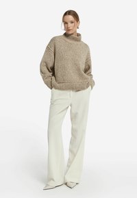 Beiger grob gestrickter Pullover mit hohem Kragen, kombiniert mit weißen, gerippten Hosen mit weitem Bein und spitzen Pumps. Strukturierter Stoff und lockere Passform.