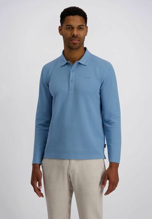 Mann steht und trägt ein hellblaues Langarm-Poloshirt und beige Hosen vor einem schlichten hellen Hintergrund.