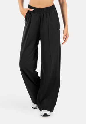 Frau trägt lockere schwarze weit geschnittene Hosen mit elastischem Bund und Seitentaschen, kombiniert mit weißen Turnschuhen, steht vor weißem Hintergrund.