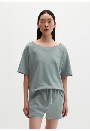 Femme portant un haut ample à manches courtes gris clair avec un large col et un short assorti avec cordon, debout devant un fond blanc uni.