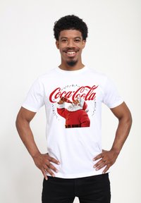 Bílý bavlněný tričko s grafikou Santa Clause pijícího Coca-Colu, s červeným nápisem a černými ozdobnými hvězdami.
