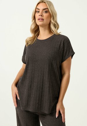 Femme blonde portant un haut à manches courtes côtelé gris foncé ample et un pantalon assorti, debout les mains dans les poches.