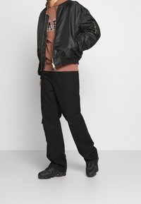 Giacca bomber nera con polsini elastici, abbinata a una felpa marrone e pantaloni cargo neri con tasche. Scarpe da ginnastica nere completano il look.