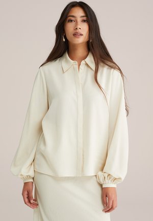 Vrouw met lang donker haar, gekleed in een crèmekleurige blouse met pofmouwen en een bijpassende rok, staand tegen een effen neutrale achtergrond.