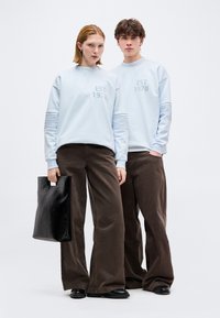 Δύο μοντέλα φορούν ανοιχτόχρωμα μπλε φούτερ με τη φράση «EST 1978», καφέ παντελόνια με φαρδιά πόδια, μαύρα παπούτσια, το ένα κρατάει μια μαύρη τσάντα tote.